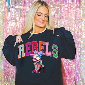 May include: A black long-sleeve sweatshirt featuring the word REBELS in large, multi-colored block letters arched across the chest. Below the text is a graphic illustration of a character wearing a colorful hat and suit, holding a cane.