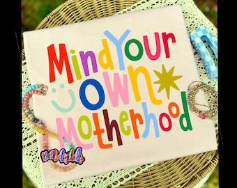 May include: A light pink t-shirt with the words "Mind Your Own Motherhood" in colorful, playful lettering. The design includes a smiley face and a star. A colorful beaded necklace and other accessories are also visible.