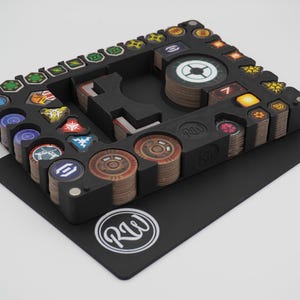 May include: Black game organiser with various compartments holding stacks of game tokens and cards. The organiser has a rectangular shape with a cutout design and is placed on a black mat. The tokens and cards have colourful designs.