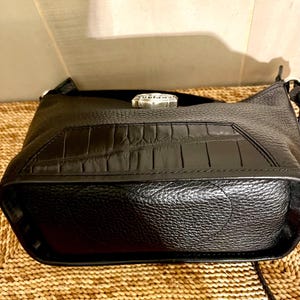 May include: A black leather handbag featuring a textured pebble grain finish on the main body and a smooth, rectangular panel with a crocodile-embossed pattern on the front. A silver-toned metal hardware piece is centered at the top edge.