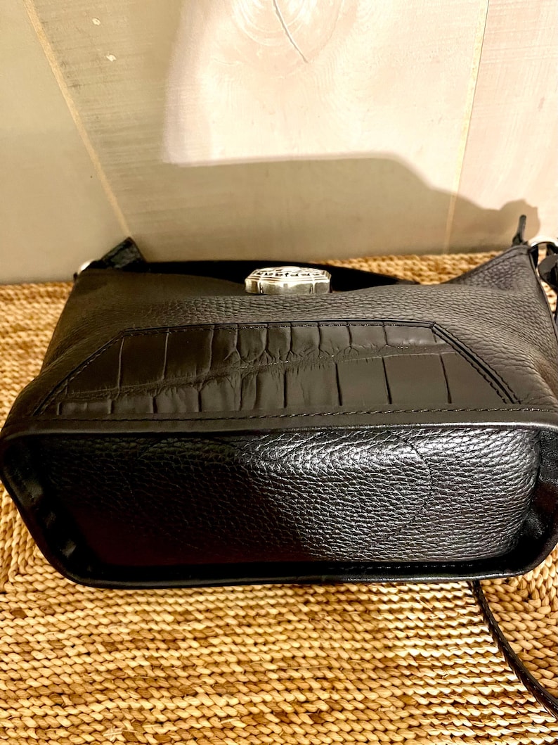 May include: A black leather handbag featuring a textured pebble grain finish on the main body and a smooth, rectangular panel with a crocodile-embossed pattern on the front. A silver-toned metal hardware piece is centered at the top edge.
