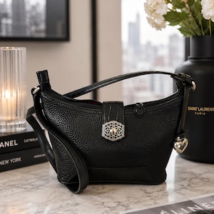 May include: A black pebbled leather handbag featuring a top zipper closure, a central vertical strap with a silver-toned ornate filigree clasp, and a small silver heart-shaped charm hanging from the side. The bag has a single shoulder strap.