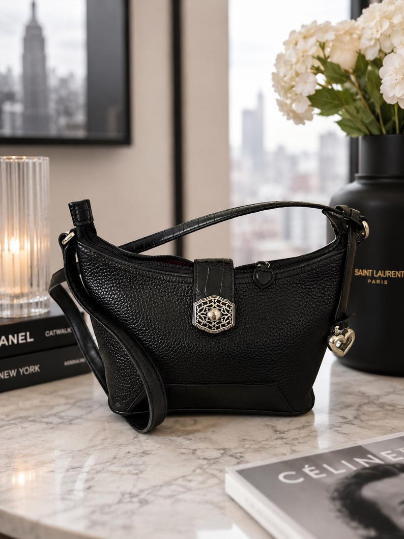 May include: A black pebbled leather handbag featuring a top zipper closure, a central vertical strap with a silver-toned ornate filigree clasp, and a small silver heart-shaped charm hanging from the side. The bag has a single shoulder strap.