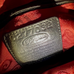 May include: A black leather patch inside a bag with red fabric lining. The patch features the embossed text Brighton, The Brighton Collection, and the serial number 8277032. The red lining has a repeating pattern of the Brighton logo throughout.