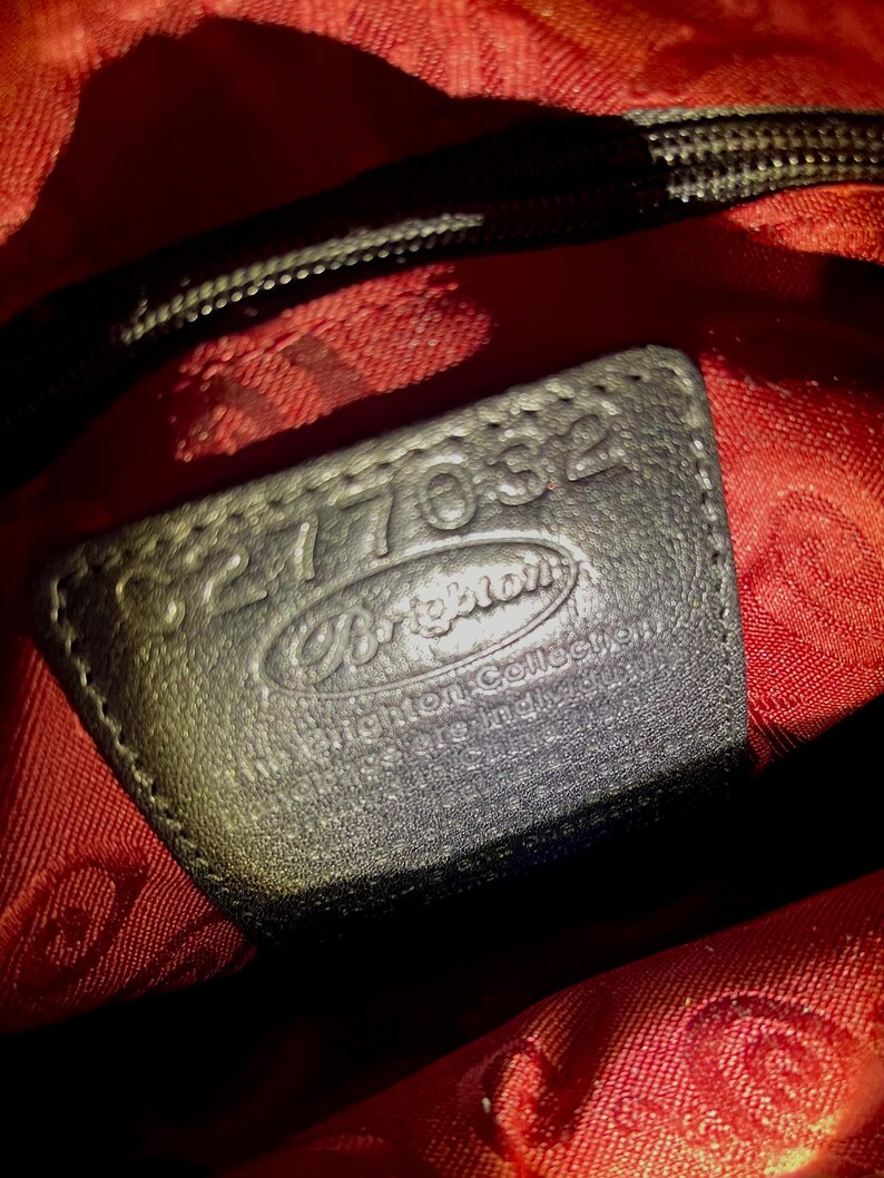 May include: A black leather patch inside a bag with red fabric lining. The patch features the embossed text Brighton, The Brighton Collection, and the serial number 8277032. The red lining has a repeating pattern of the Brighton logo throughout.
