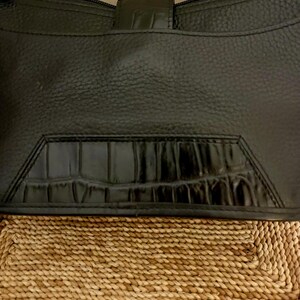 May include: A black handbag with a pebbled texture and a smooth, glossy crocodile-embossed leather panel at the bottom. The bag features a top zipper closure, a thin shoulder strap, and a central vertical tab detail with a matching crocodile finish.