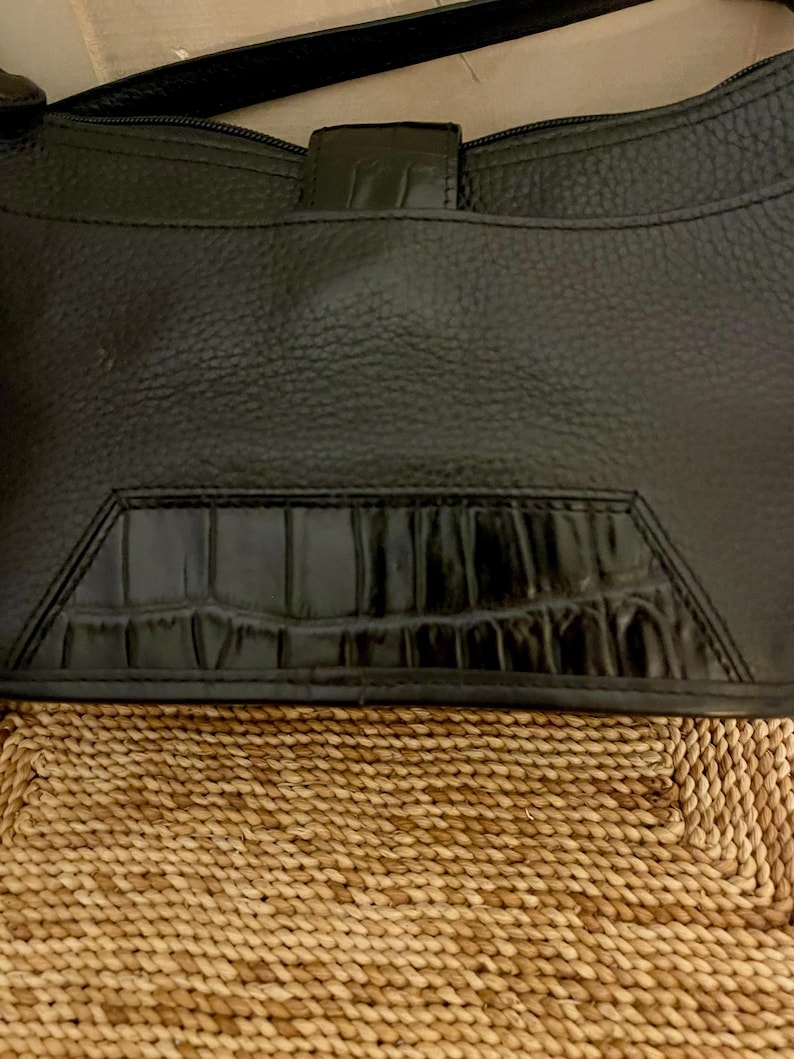 May include: A black handbag with a pebbled texture and a smooth, glossy crocodile-embossed leather panel at the bottom. The bag features a top zipper closure, a thin shoulder strap, and a central vertical tab detail with a matching crocodile finish.