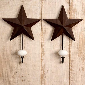 Puede incluir: Dos colgadores de pared rústicos en forma de estrella de metal marrón oscuro. Cada estrella está montada sobre una varilla negra con un acento blanco ovalado y un pequeño gancho. Los colgadores se muestran sobre un fondo de madera clara y envejecida.