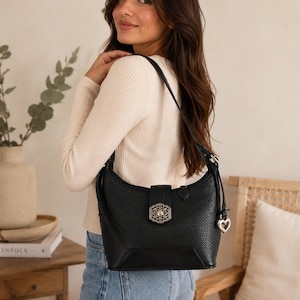 May include: A black pebbled leather shoulder bag featuring a silver-toned filigree metal clasp on the front flap and a matching silver heart-shaped charm hanging from the side strap. The bag has a structured base and a single adjustable shoulder strap.