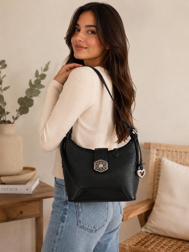 May include: A black pebbled leather shoulder bag featuring a silver-toned filigree metal clasp on the front flap and a matching silver heart-shaped charm hanging from the side strap. The bag has a structured base and a single adjustable shoulder strap.