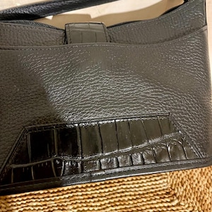 May include: A black handbag featuring a textured pebbled leather body and a smooth, glossy panel with a crocodile-embossed pattern at the base. The bag includes a top zipper closure, a matching black shoulder strap, and a central vertical tab.