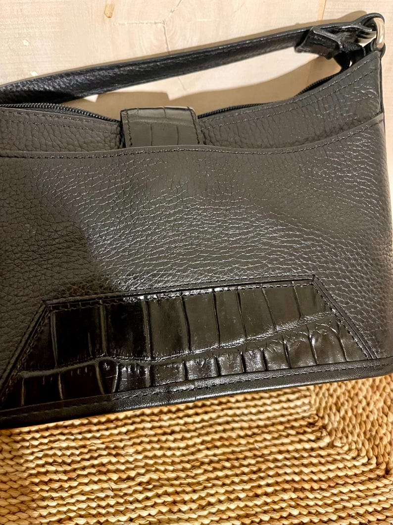 May include: A black handbag featuring a textured pebbled leather body and a smooth, glossy panel with a crocodile-embossed pattern at the base. The bag includes a top zipper closure, a matching black shoulder strap, and a central vertical tab.