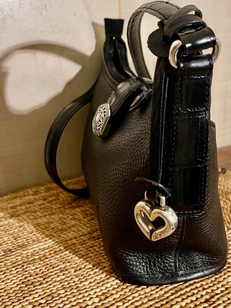 May include: A black leather handbag with a pebbled texture, featuring a smooth crocodile-embossed leather strap and trim. The bag is adorned with a silver-toned heart-shaped charm and a decorative circular metal ornament on the side.