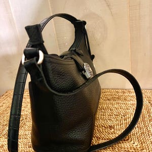May include: A black pebbled leather handbag featuring a structured silhouette, a single shoulder strap with a subtle crocodile-embossed texture, and a silver-toned metal decorative emblem attached to the front with a vertical leather strap.