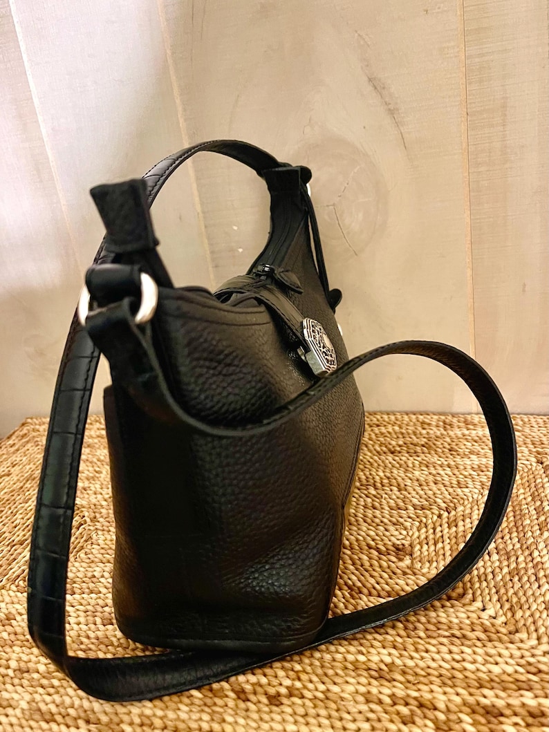 May include: A black pebbled leather handbag featuring a structured silhouette, a single shoulder strap with a subtle crocodile-embossed texture, and a silver-toned metal decorative emblem attached to the front with a vertical leather strap.