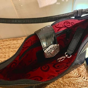 May include: A black handbag with a textured exterior and a strap. The interior is lined with red fabric featuring a black swirling pattern. A black flap with a silver-toned metal filigree clasp secures the opening, and a metal clip is attached.
