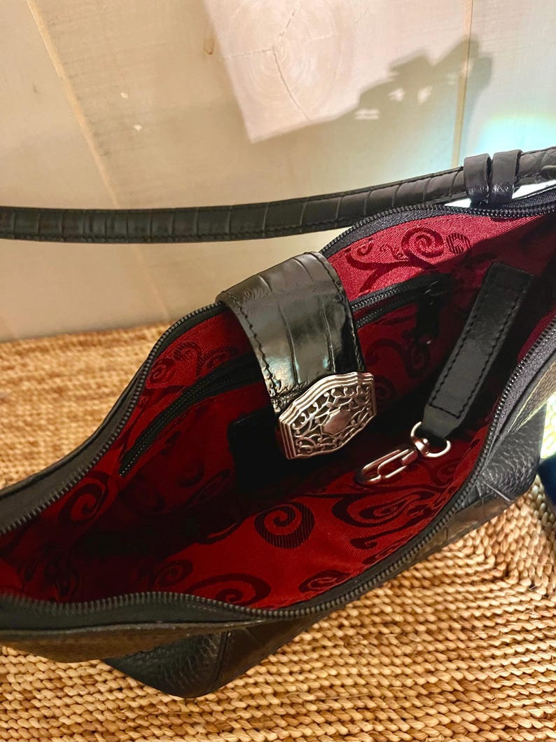 May include: A black handbag with a textured exterior and a strap. The interior is lined with red fabric featuring a black swirling pattern. A black flap with a silver-toned metal filigree clasp secures the opening, and a metal clip is attached.