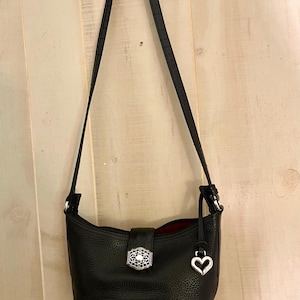 May include: A black leather shoulder bag with a pebbled texture and a long adjustable strap. The bag features a central silver-toned decorative clasp with intricate filigree detailing and a dangling silver heart-shaped charm attached to the side.