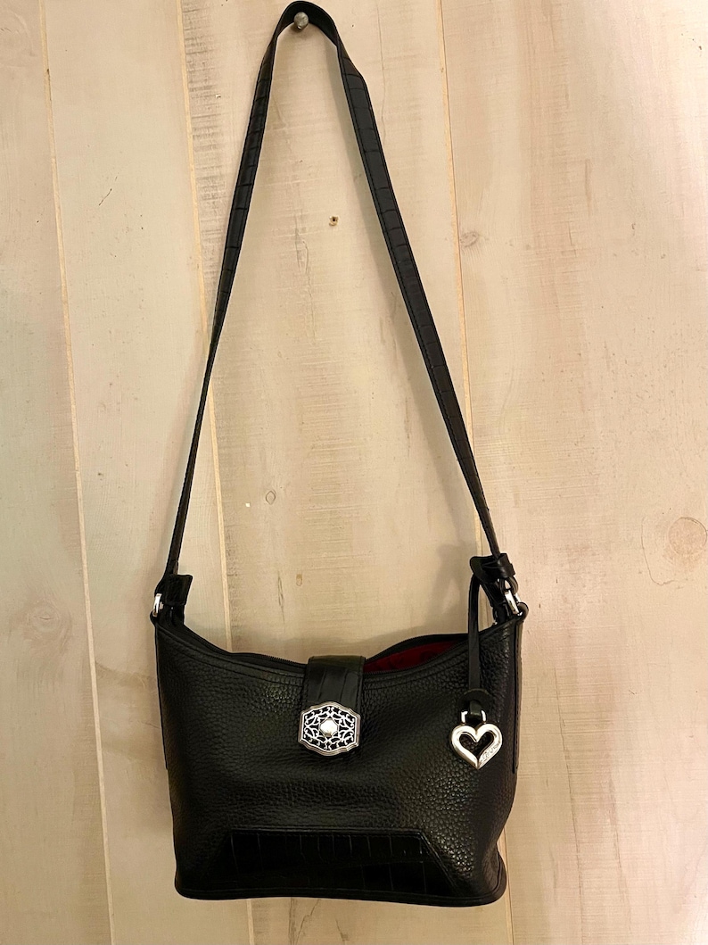 May include: A black leather shoulder bag with a pebbled texture and a long adjustable strap. The bag features a central silver-toned decorative clasp with intricate filigree detailing and a dangling silver heart-shaped charm attached to the side.
