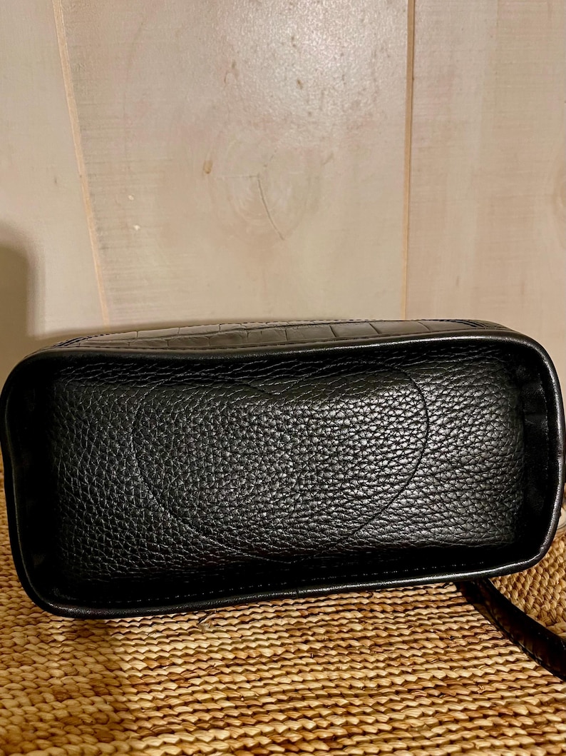 May include: A black rectangular bag with a textured surface featuring a prominent embossed heart shape on the front panel. The bag has a structured frame with smooth edges and a distinct crocodile-like pattern along the top border.