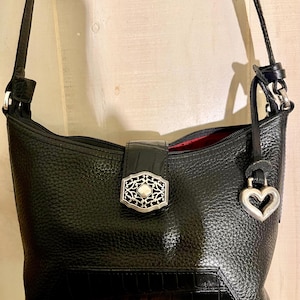 May include: A black shoulder bag with a pebbled texture and a smooth crocodile-embossed panel at the base. It features a central magnetic closure with an ornate silver-tone metal emblem, a matching silver heart-shaped charm, and a red interior lining.