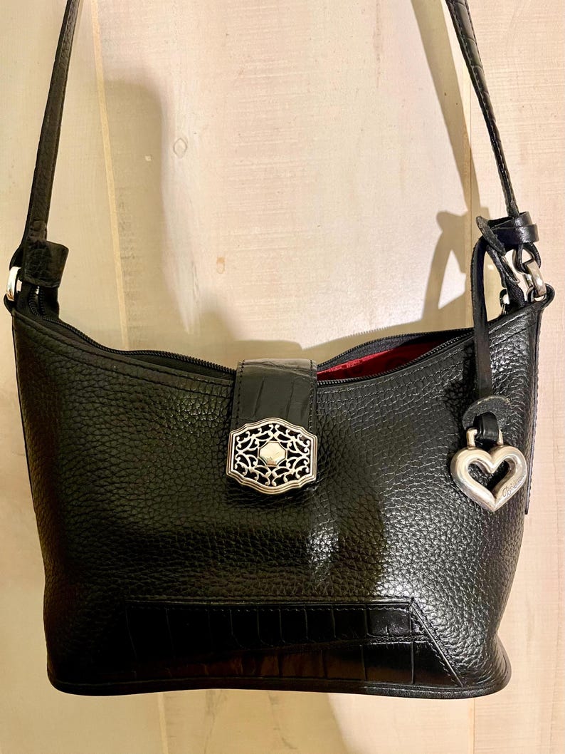 May include: A black shoulder bag with a pebbled texture and a smooth crocodile-embossed panel at the base. It features a central magnetic closure with an ornate silver-tone metal emblem, a matching silver heart-shaped charm, and a red interior lining.