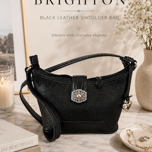 May include: A black leather shoulder bag with a textured surface, featuring a central silver-toned decorative clasp with an intricate filigree pattern, a top zipper closure with a heart-shaped pull, and a dangling silver heart charm on the side.