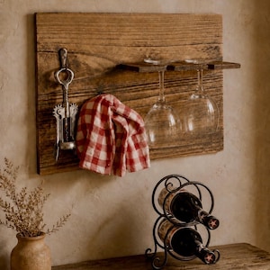 May include: A wall-mounted wooden rack featuring a metal corkscrew, a red and white checkered cloth, and two hanging glass wine stems. Below, a black metal wine rack holds two bottles, accompanied by a small tan vase with dried foliage.