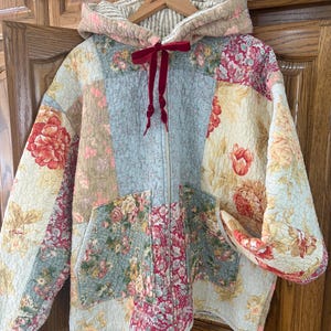 May include: A quilted zip-up hoodie featuring a patchwork design of various floral patterns in shades of red, yellow, blue, and cream. The garment includes a hood with a striped interior lining and a dark red velvet ribbon drawstring at the neck.