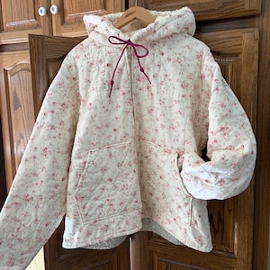 May include: A cream-colored quilted zip-up hoodie featuring a delicate pink and red floral pattern throughout. The garment includes a hood with a bright magenta drawstring, long sleeves, and two front patch pockets. The fabric has a stitched texture.