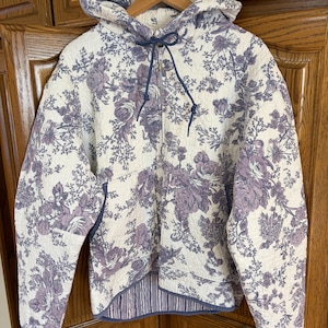 Könnte beinhalten: Gesteppter Kapuzenpullover mit floralem Muster in Lila und Weiß. Die Jacke hat einen Reißverschluss, eine Kapuze mit Kordelzug und ein gestreiftes Futter. Das Blumenmuster zeigt Rosen und andere Blumen. Bequemer, lässiger Stil.
