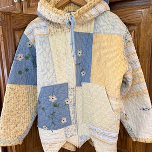 May include: A quilted jacket with a patchwork design in shades of yellow, blue, and white. The jacket features a hood, a front zipper, and two pockets. Floral embroidery accents the blue patches. The jacket is hanging on a wooden hanger.