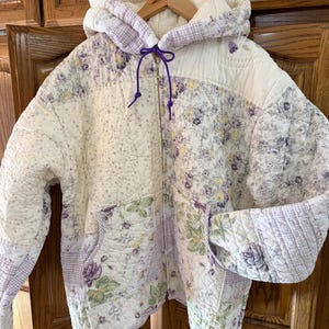 Puede incluir: Una sudadera acolchada con cremallera y capucha que presenta un diseño de patchwork con patrones florales en crema, lavanda y amarillo. La prenda incluye una capucha con cordón morado, mangas largas y dos bolsillos frontales de tela cosida.