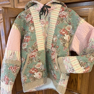May include: Quilted hoodie with a floral pattern in shades of green, pink, and beige. The jacket features a drawstring hood, front pockets, and patchwork detailing on the sleeves and body. The overall design is a mix of vintage and bohemian styles.