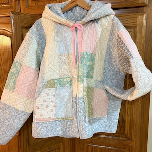 May include: A quilted zip-up hooded jacket featuring a patchwork design of various floral and gingham patterns in soft pastel shades of blue, pink, green, and white. The garment includes two front pockets and a bright pink drawstring at the neck.