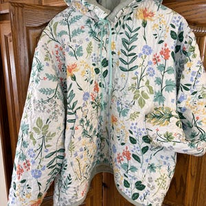 May include: A quilted jacket with a floral pattern in shades of white, green, yellow, orange, and blue. The jacket has a hood and a light green lining. It features a zipper closure and two front pockets. The jacket is hanging on a wooden hanger.