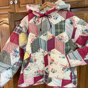 Puede incluir: Una chaqueta acolchada con capucha que presenta un diseño de patchwork con diversas telas, incluyendo estampados florales, cuadros vichy rojos y blancos, cuadros amarillos y cuadros verdes. Tiene cremallera frontal, dos bolsillos y cordón.