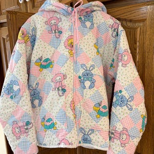 May include: A quilted jacket with a hood, featuring a patchwork design of pink, blue, and white squares. The jacket is adorned with cartoon animals, including monkeys, bunnies, and turtles. It has a full zipper and a pink drawstring.
