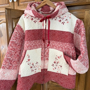 May include: A quilted zip-up hooded jacket featuring a patchwork design in shades of red, pink, and cream. The jacket includes two front pockets with embroidered floral vines, a matching hood, and a dark red ribbon tie at the neckline.