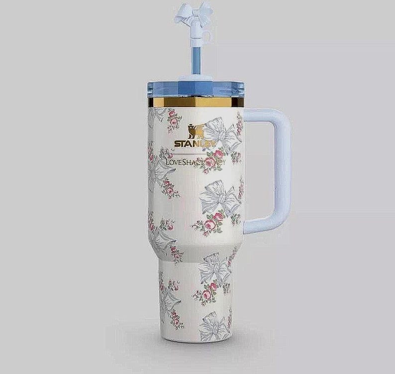 K&ouml;nnte beinhalten: Ein wei&szlig;er Stanley-Becher mit einem Blumendesign aus Schleifen und Rosen. Er hat einen hellblauen Griff, Deckel und Strohhalm mit einer Schleife. Der Text "STANLEY LOVESHACK" ist auf der Seite aufgedruckt.