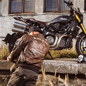 Op de afbeelding: Bruine leren bomberjack met de tekst "BLACK HEART MOTORCYCLE GEAR" en een motorfietsafbeelding op de rug. Een zwarte motorfiets staat in de buurt geparkeerd. De motorfiets heeft een zwart frame, bruin zadel en gouden voorvorken.