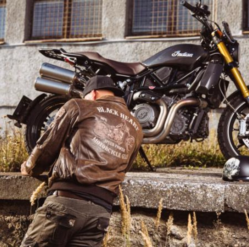 Op de afbeelding: Bruine leren bomberjack met de tekst "BLACK HEART MOTORCYCLE GEAR" en een motorfietsafbeelding op de rug. Een zwarte motorfiets staat in de buurt geparkeerd. De motorfiets heeft een zwart frame, bruin zadel en gouden voorvorken.