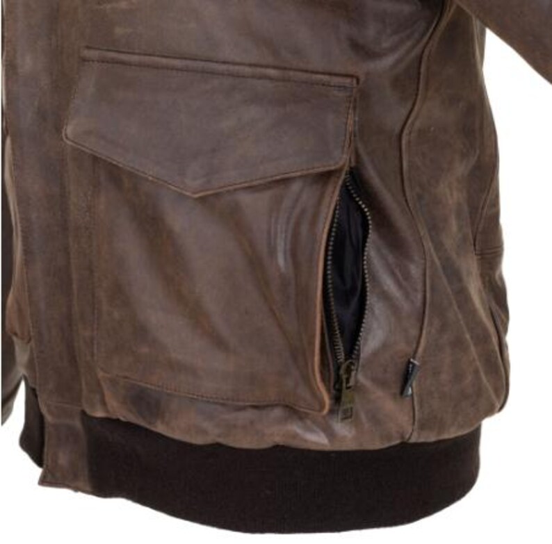 Op de afbeelding: Close-up van een bruine leren jas met een zak en een rits. De jas heeft een distressed look, met zichtbare slijtage. De onderkant van de jas heeft een donkerbruine geribbelde zoom.