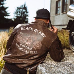 Op de afbeelding: Bruine leren jas met een motorfietsafbeelding op de rug. De jas heeft de woorden "BLACK HEART MOTORCYCLE GEAR" in witte letters. Een zwarte pet wordt gedragen.