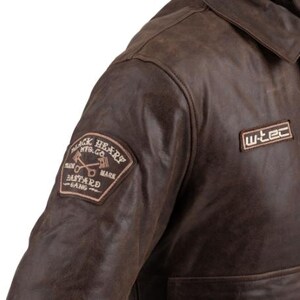 Op de afbeelding: Bruine leren jas met kraag en twee patches. E&eacute;n patch luidt "BLACK HEART MFG. CO. PLAIN BASTARD GANG MARK" en de andere "w-tec". De jas heeft een verweerde uitstraling.