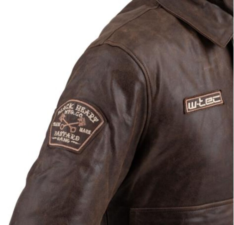 Op de afbeelding: Bruine leren jas met kraag en twee patches. E&eacute;n patch luidt "BLACK HEART MFG. CO. PLAIN BASTARD GANG MARK" en de andere "w-tec". De jas heeft een verweerde uitstraling.