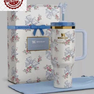 K&ouml;nnte beinhalten: Ein wei&szlig;er Stanley Loveshackfancy Becher mit hellblauem Griff und Deckel, verziert mit einem Blumen- und Schleifenmuster. Er wird mit einer passenden Geschenkbox und einem blauen Tuch pr&auml;sentiert.