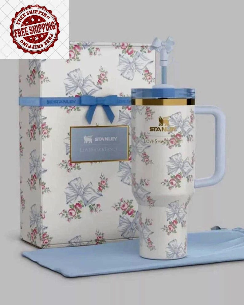 K&ouml;nnte beinhalten: Ein wei&szlig;er Stanley Loveshackfancy Becher mit hellblauem Griff und Deckel, verziert mit einem Blumen- und Schleifenmuster. Er wird mit einer passenden Geschenkbox und einem blauen Tuch pr&auml;sentiert.