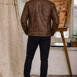 Op de afbeelding: Bruine leren jas met een klassiek design, kraag en knopen. De jas wordt gecombineerd met een zwarte jeans en bruine laarzen. De persoon staat in een kamer met een neutrale achtergrond.