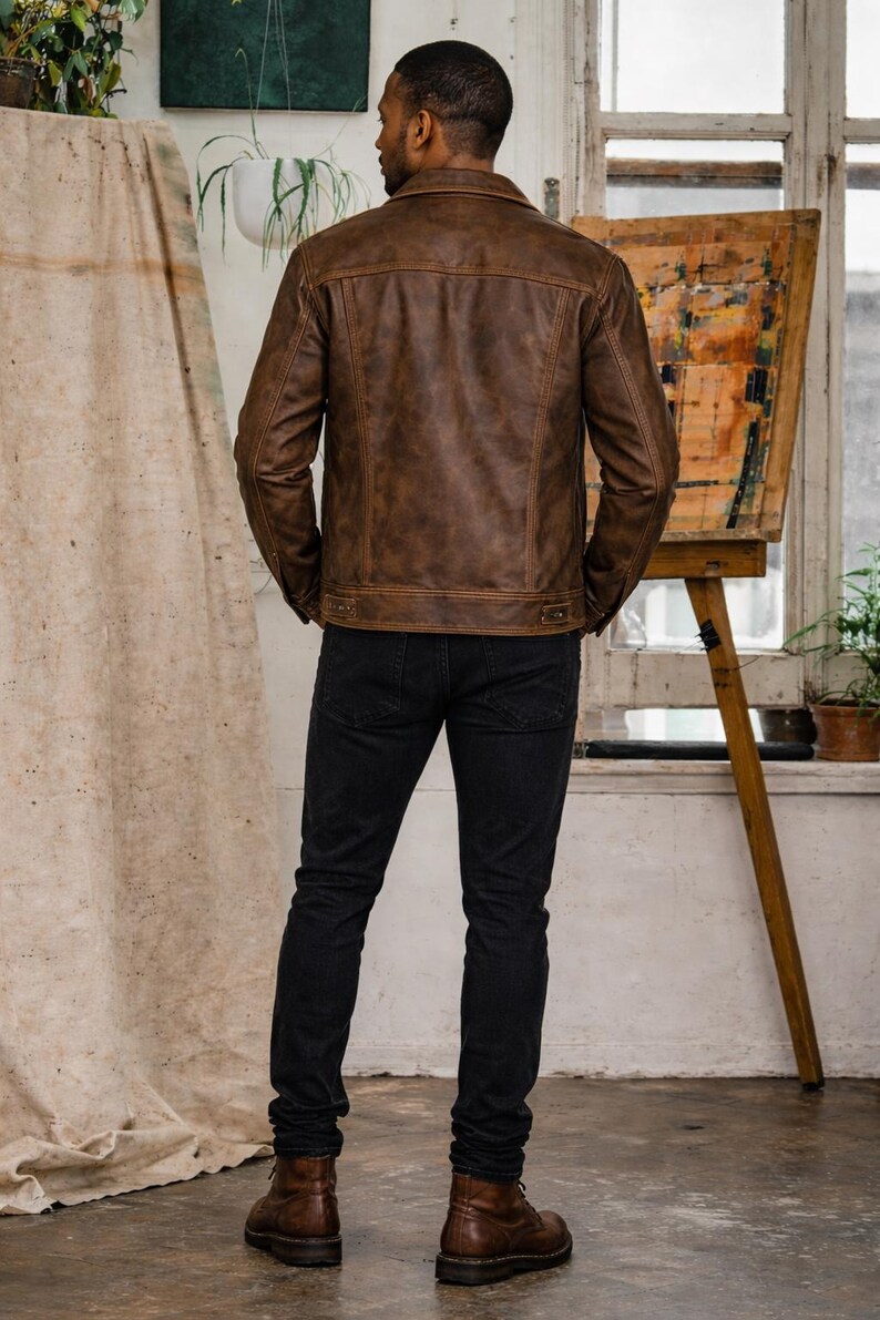 Op de afbeelding: Bruine leren jas met een klassiek design, kraag en knopen. De jas wordt gecombineerd met een zwarte jeans en bruine laarzen. De persoon staat in een kamer met een neutrale achtergrond.